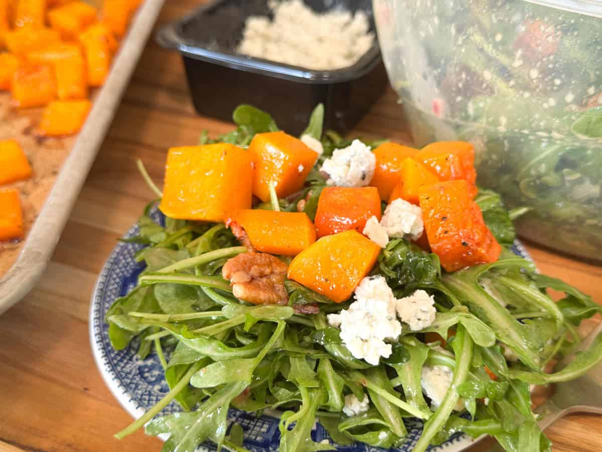 A salad on a plate, featuring roasted butternut squash cubes, crumbled cheese, and walnuts on a bed of arugula. In the background, a tray with more roasted squash and a container of salad are visible.