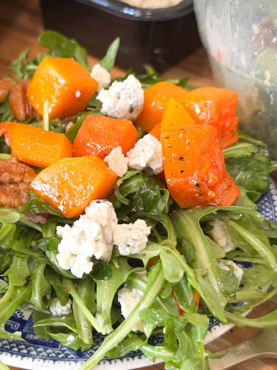 A salad featuring roasted orange squash cubes, arugula, crumbled blue cheese, and pecans is arranged on a blue-patterned plate. The dish is fresh and colorful, with textures ranging from creamy cheese to crunchy pecans.