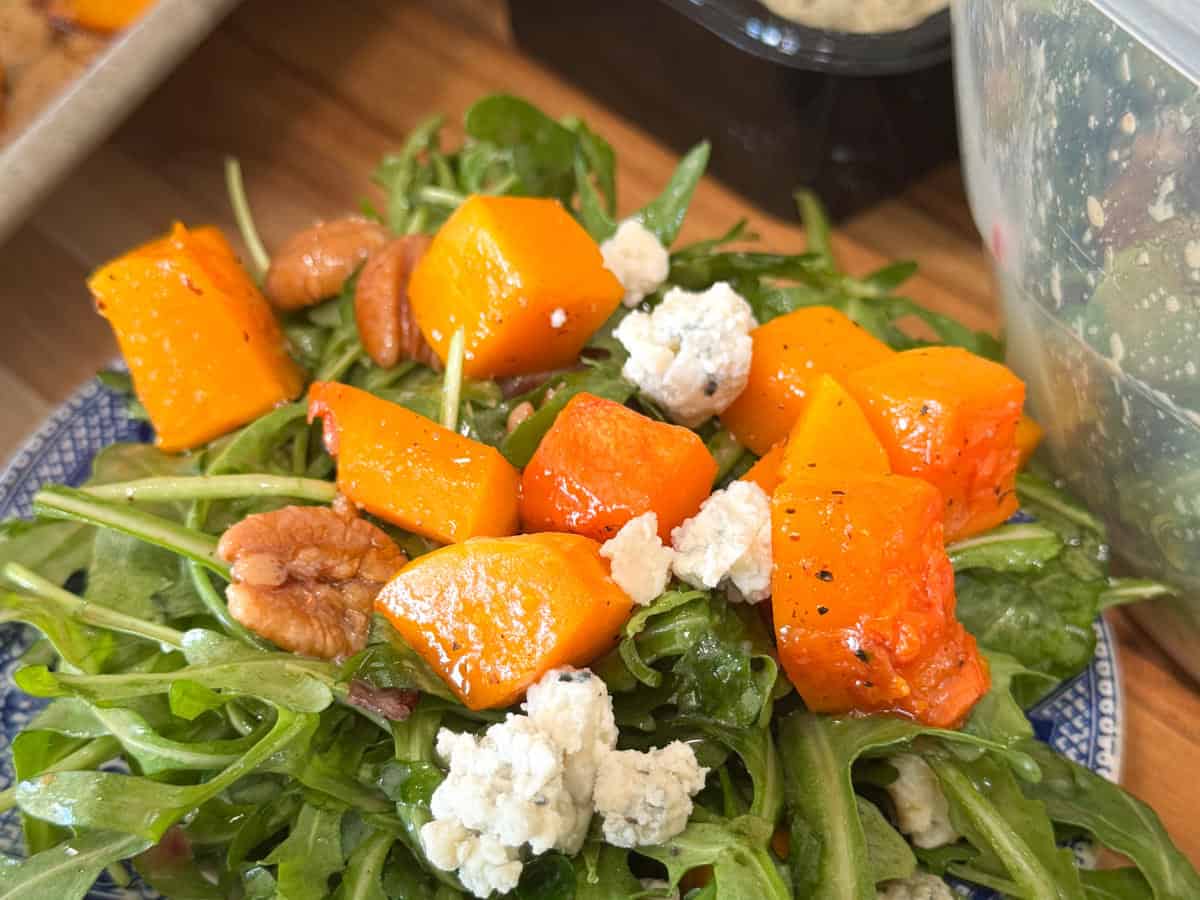 A salad with roasted orange pumpkin cubes, crumbled blue cheese, and pecans on a bed of arugula, served on a blue and white plate. A plastic container and a black dish are partially visible in the background.
