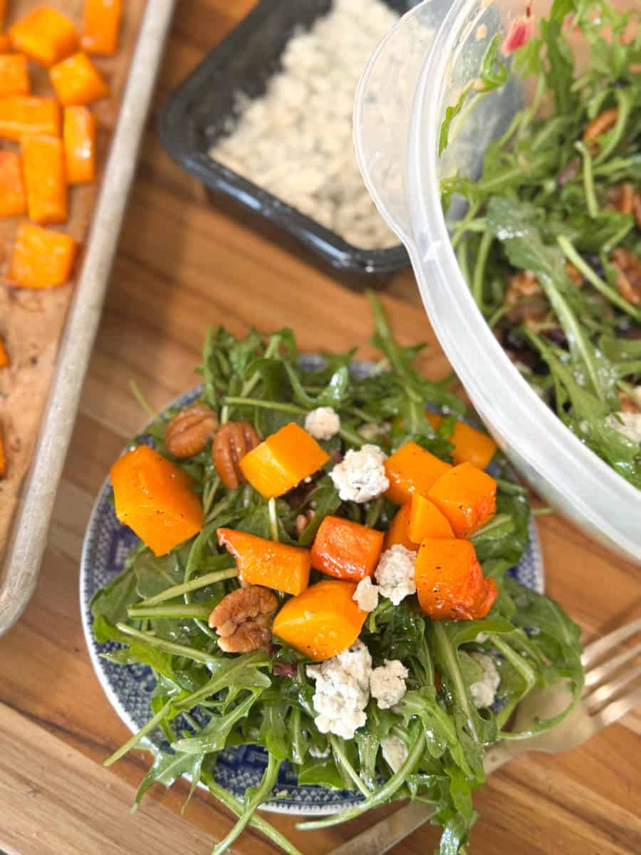 A salad with arugula, roasted butternut squash, pecans, and blue cheese crumbles is served on a plate. A tray of roasted squash and a container of the salad mix are nearby on a wooden surface.