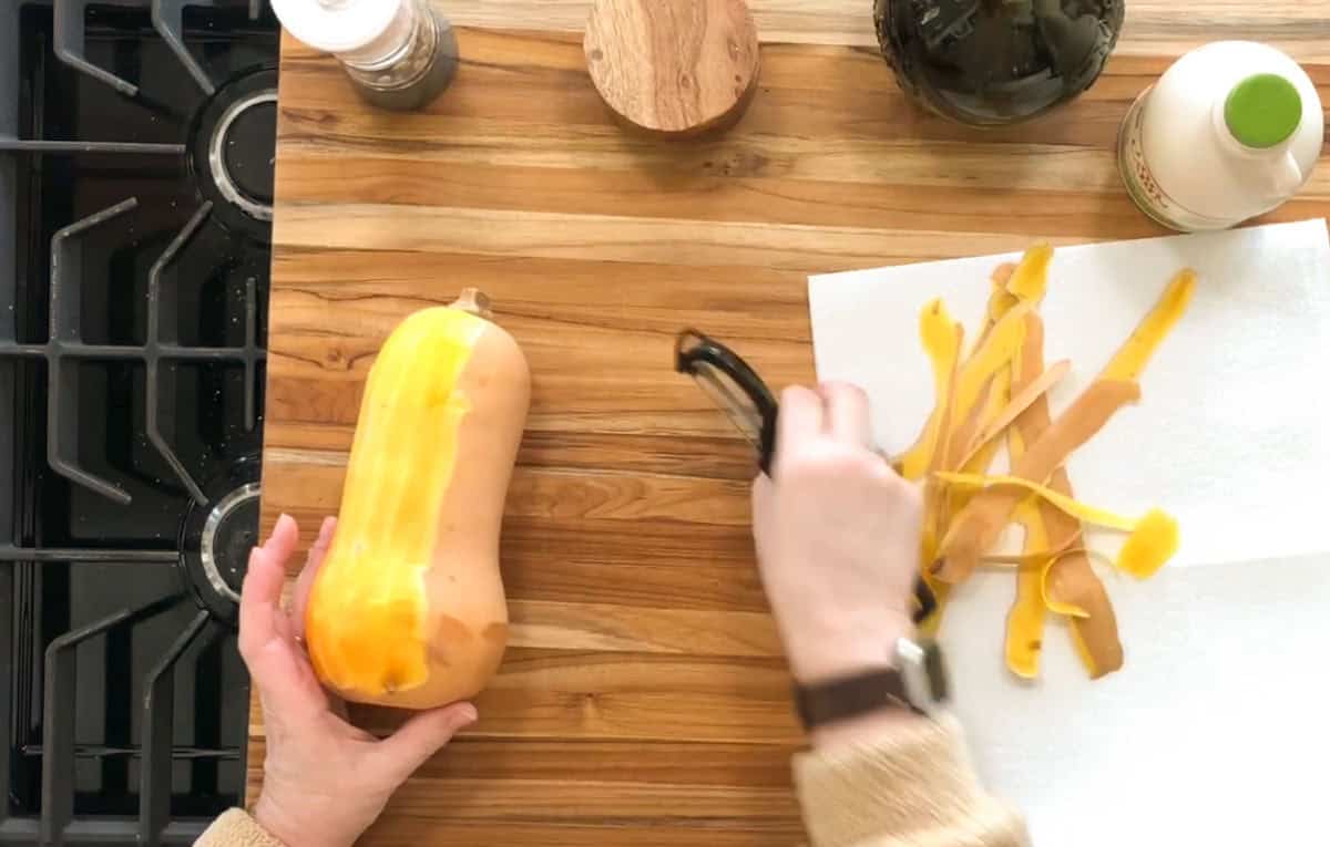 A person is peeling a butternut squash on a wooden countertop. The squash is held in one hand, while the other hand uses a peeler. Strips of squash peel are on a paper towel. Kitchen items, like a pepper grinder and bottles, are nearby.