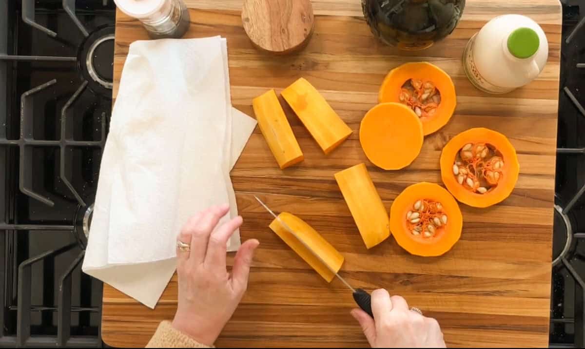 A person is slicing butternut squash on a wooden cutting board. The squash is halved, with seeds visible in one half. A bottle, a bowl, and paper towels are nearby. The person holds a slice while cutting with a knife.