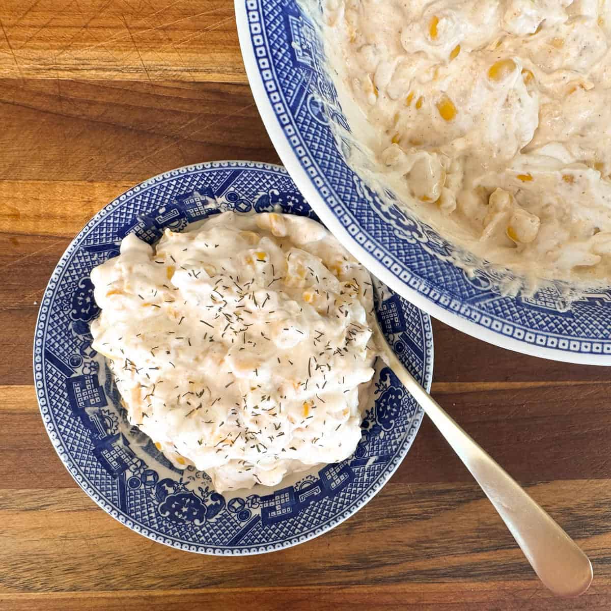 Two blue-patterned bowls contain creamy corn dip. The small bowl has a serving with a sprinkle of dill on top and a spoon. The larger bowl is filled with the dip, sitting on a wooden surface.
