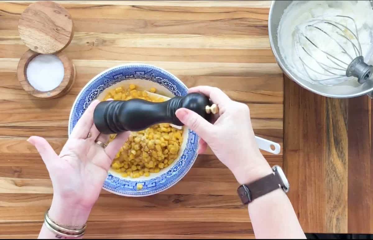 A person holds a black pepper grinder over a bowl of food, likely corn, on a wooden countertop. Nearby, there is a small bowl of salt and a mixing bowl with a whisk attachment resting inside it.