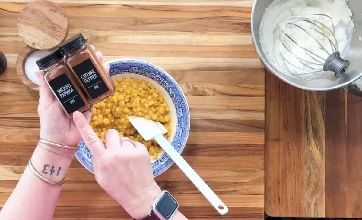 Hands holding jars of smoked paprika and cayenne pepper over a bowl of corn with a spatula in it. A mixing bowl with a whisk is on the right. Both hands have jewelry and the left wrist has a smartwatch. Wooden countertop background.