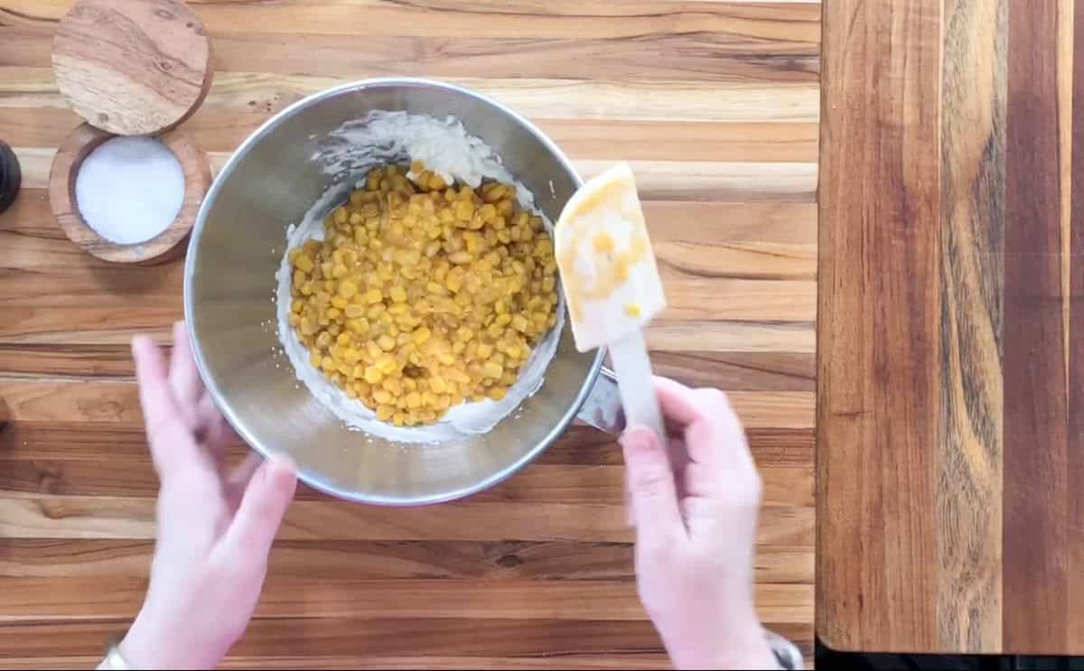 Hands hold a metal mixing bowl with corn and batter mixture. A spatula rests inside the bowl. A small wooden container with salt is visible on the wooden countertop. An empty wooden board is on the right.