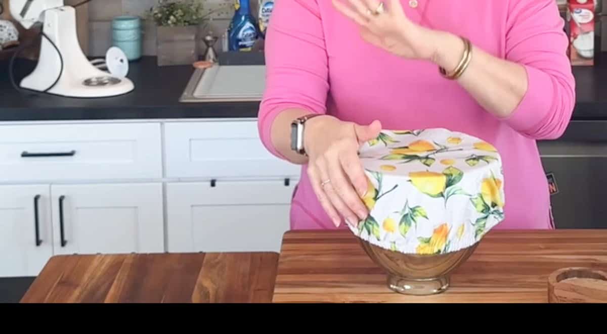 A person in a pink shirt places a yellow floral cover over a bowl on a wooden kitchen counter. The background includes a sink, a white mixer, and various kitchen items.