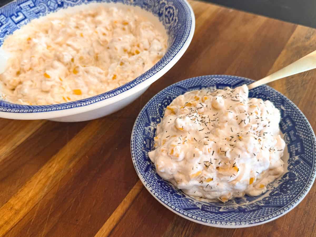 Two bowls on a wooden surface: a larger bowl filled with creamy corn dip and a smaller blue-patterned bowl containing a serving of the dip. The smaller bowl has a spoon and is garnished with dried herbs.
