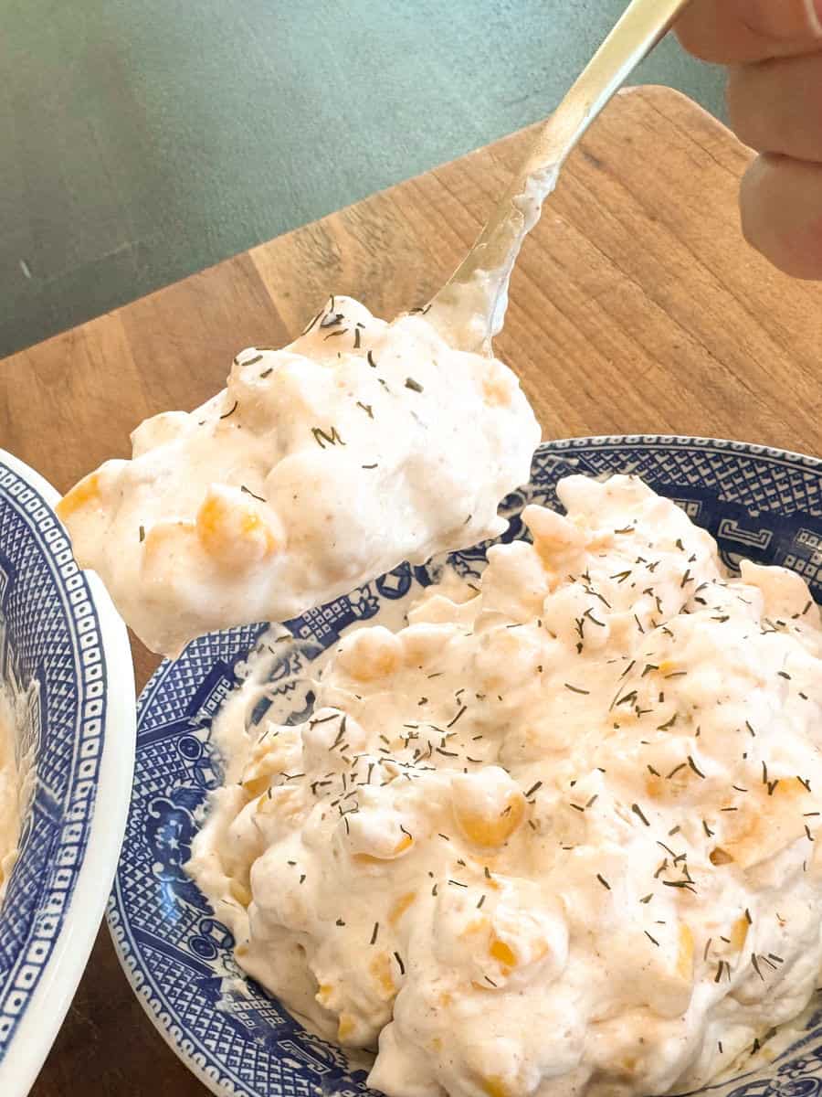 A person holds a spoonful of a creamy mixture over a blue and white bowl filled with the same dish. The mixture includes corn and is sprinkled with herbs, likely dill. The scene is set on a wooden table.