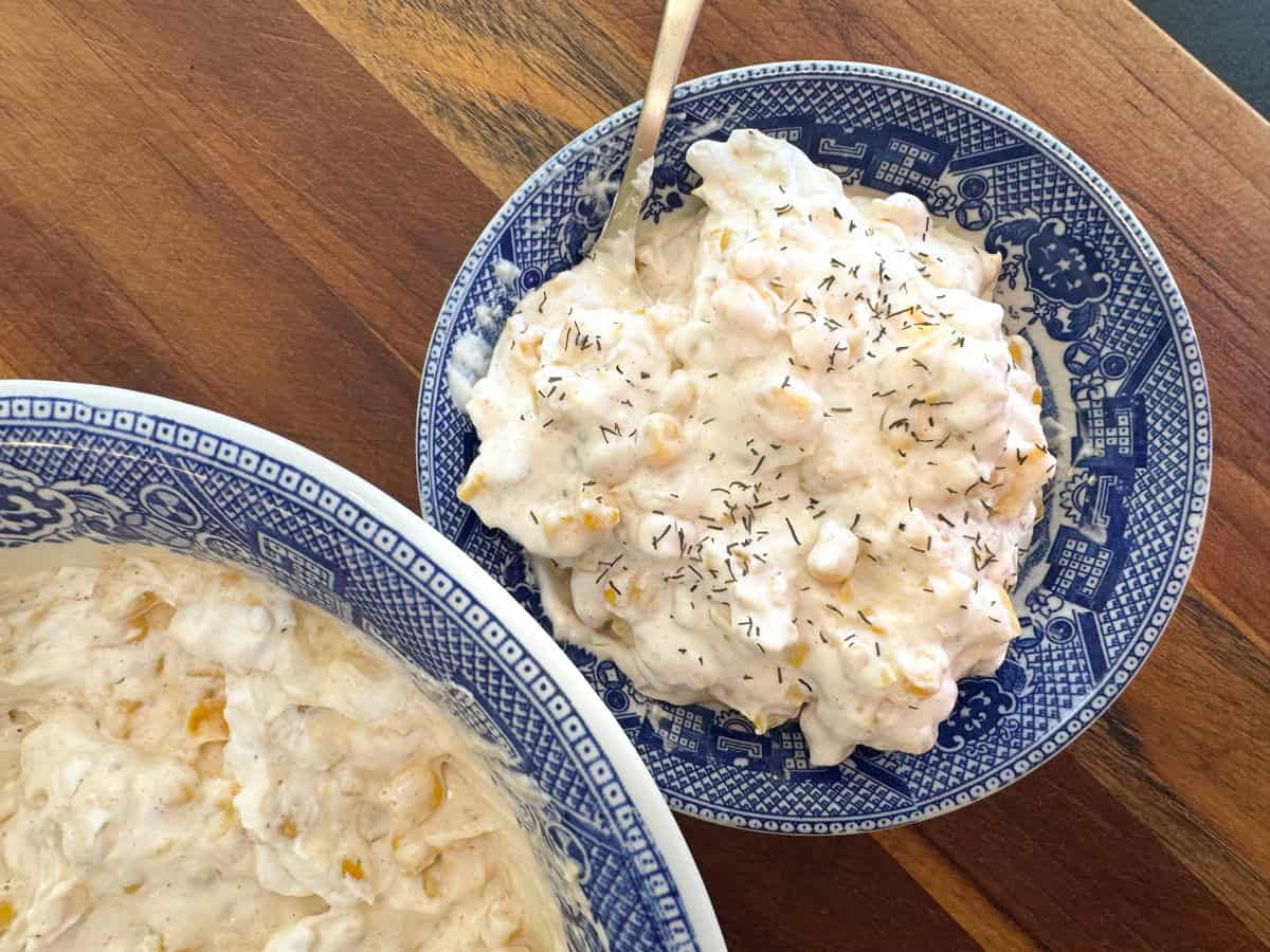 A blue patterned bowl filled with a creamy salad sits on a wooden table. The salad includes corn and is sprinkled with dried herbs. A silver spoon rests in the bowl. Another dish containing more of the salad is partially visible.