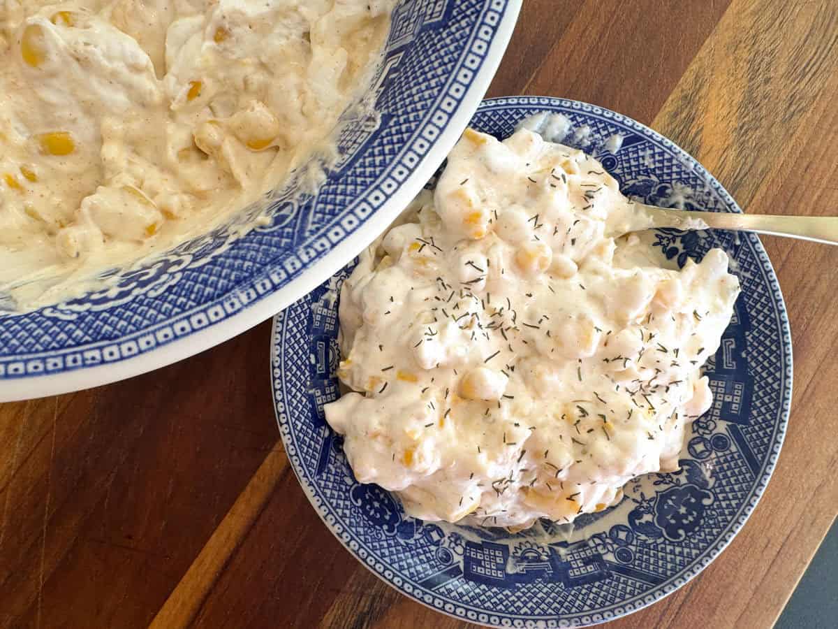 A serving of creamy corn dip topped with herbs on a blue patterned plate, next to a larger bowl of the same dip, placed on a wooden surface.