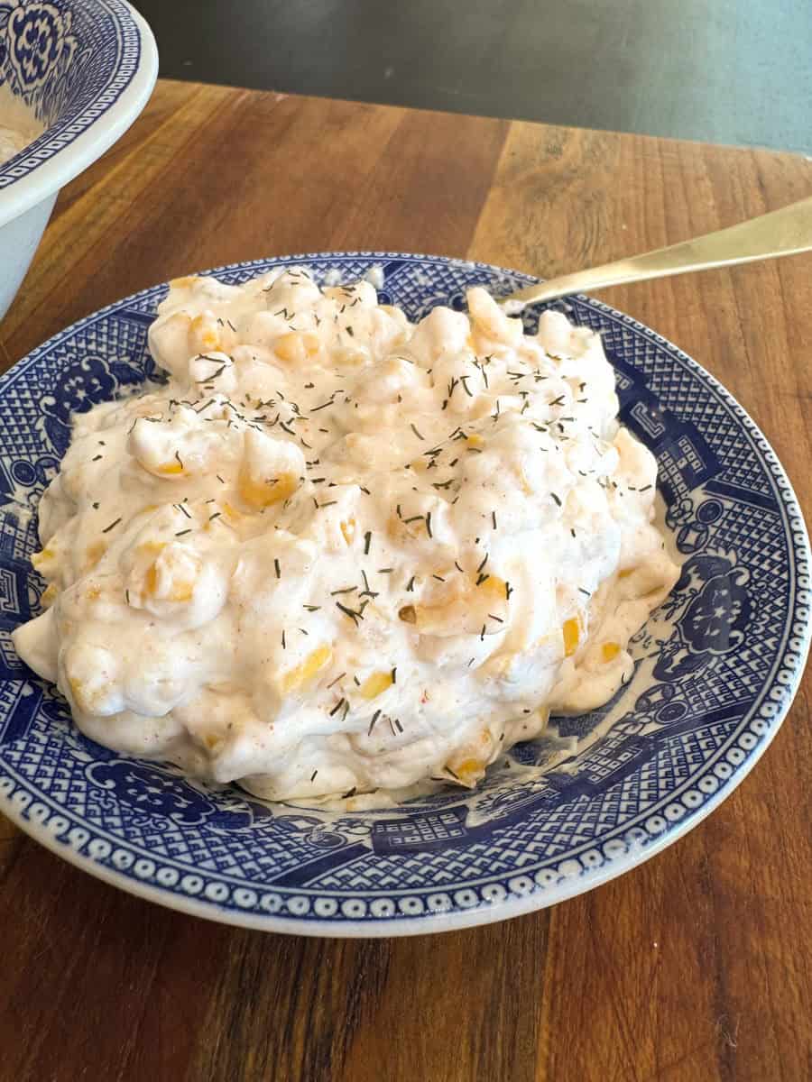 A blue and white patterned bowl filled with a creamy pasta salad. The salad consists of pasta shells, small corn kernels, and is topped with a sprinkling of herbs. It is placed on a wooden table.
