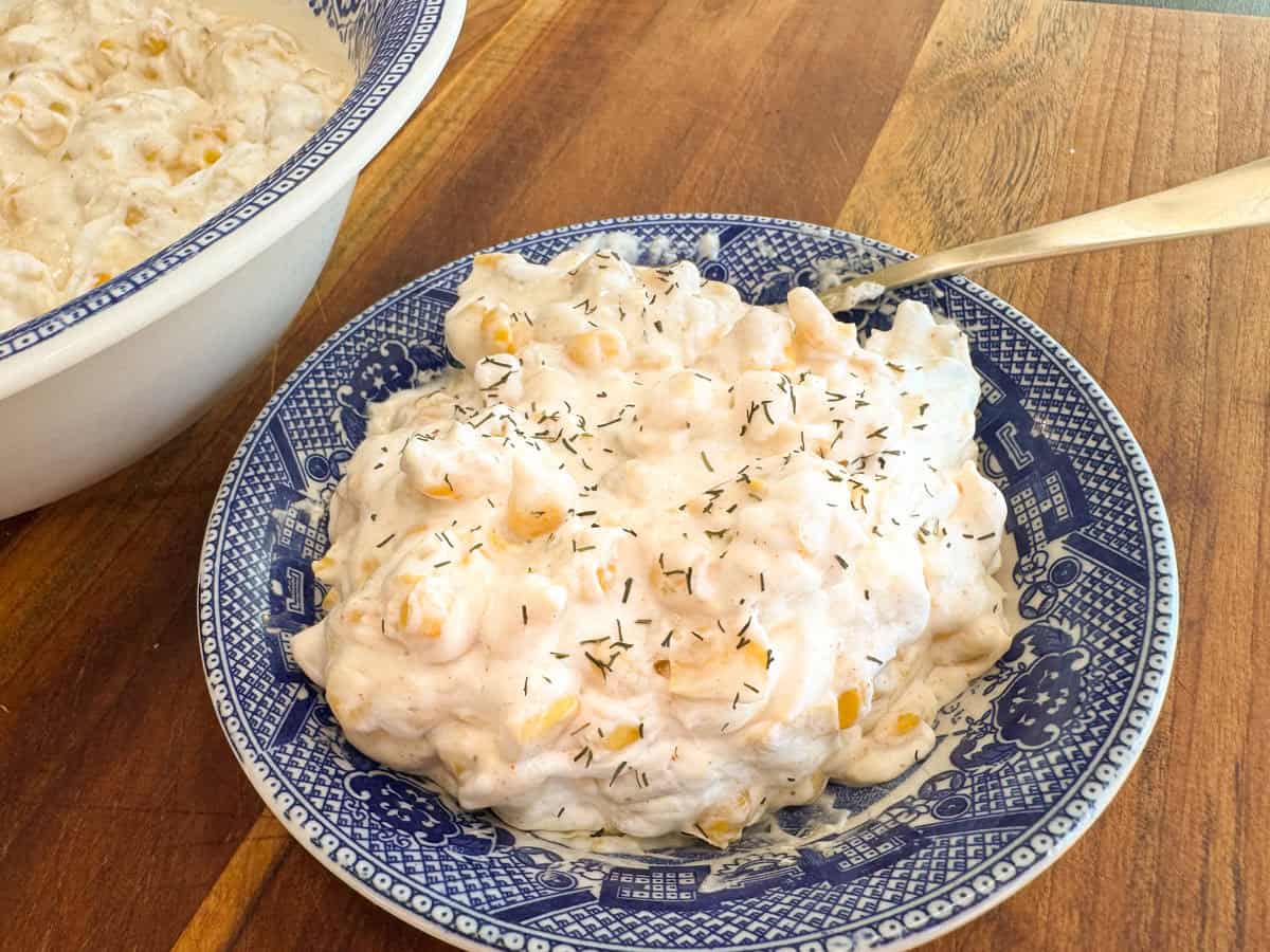 A blue and white patterned bowl filled with creamy corn salad, garnished with herbs, on a wooden surface. A spoon is placed in the salad. Part of a larger, similar bowl is visible in the background.