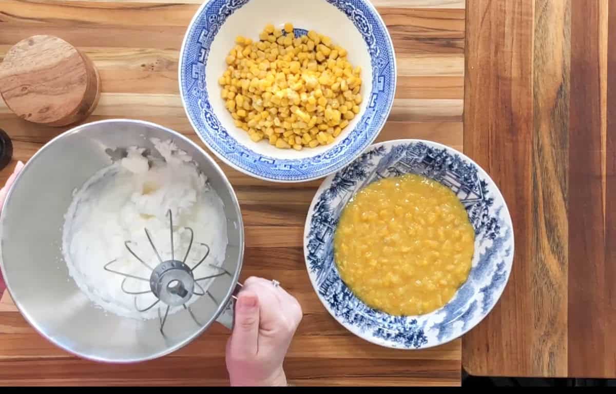 A person is using a mixer with cream in a metal bowl. Nearby, there are two bowls: one with corn and the other with a corn mixture. Everything is set on a wooden table, alongside a small wooden container.
