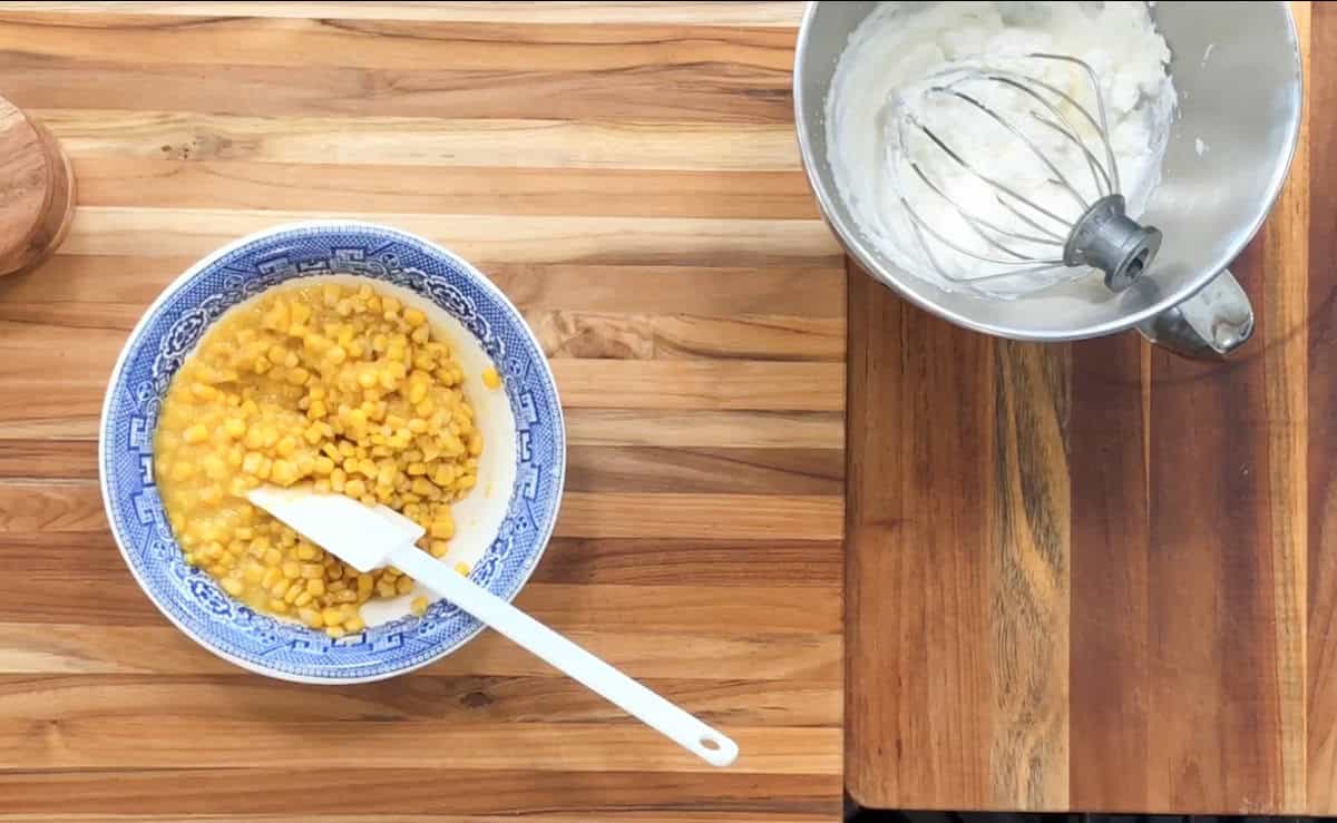 A blue and white bowl filled with corn sits on a wooden countertop. A white spatula rests inside the bowl. Next to it is a metal mixing bowl with a whisk attachment, containing a white creamy substance.