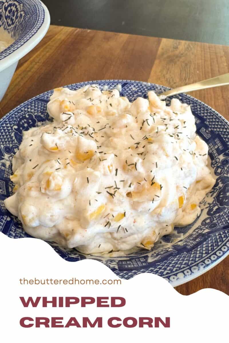 A blue and white plate filled with whipped cream corn topped with herbs. The plate is on a wooden surface. The text on the image reads "thebutteredhome.com Whipped Cream Corn.