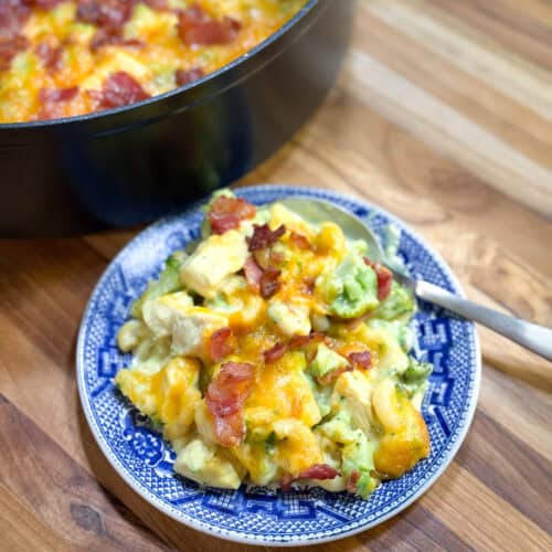 A plate with a serving of cheesy chicken, broccoli, and bacon casserole on a blue and white dish, with a spoon beside it and the casserole dish in the background on a wooden surface.