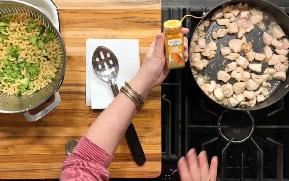 A person holds a container of turmeric over a stovetop skillet with diced chicken; beside it, cooked pasta with broccoli sits in a colander on a wooden cutting board with a slotted spoon and paper towels.