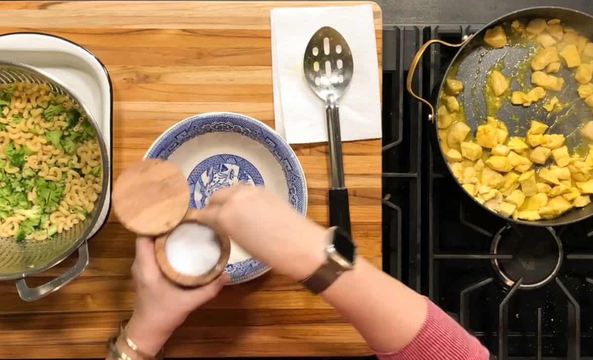 Overhead view of a person adding salt to a blue and white bowl on a wooden counter, with cooked macaroni and broccoli on the left and a pan of cooked chicken pieces on the stove to the right.