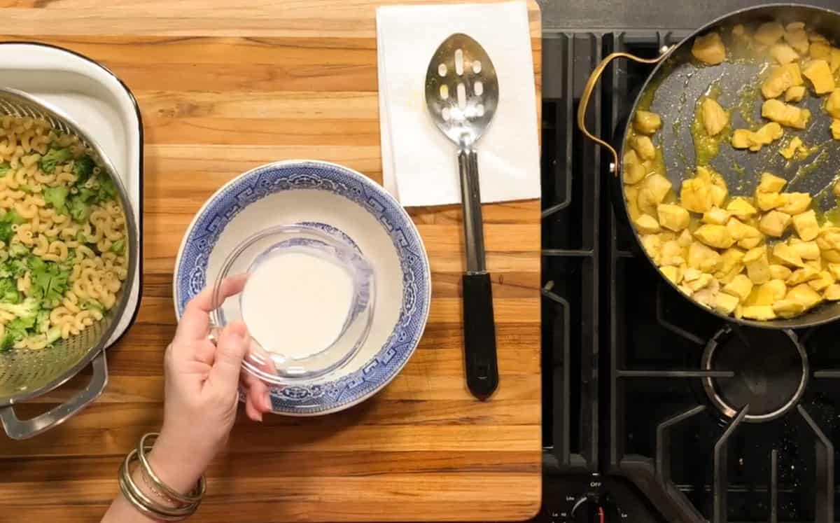 A hand holds a small glass bowl of milk above a blue-patterned bowl. Cooked pasta with broccoli is in a colander on the left, and a pan with cooked chicken is on the stove to the right. A slotted spoon rests on a napkin.
