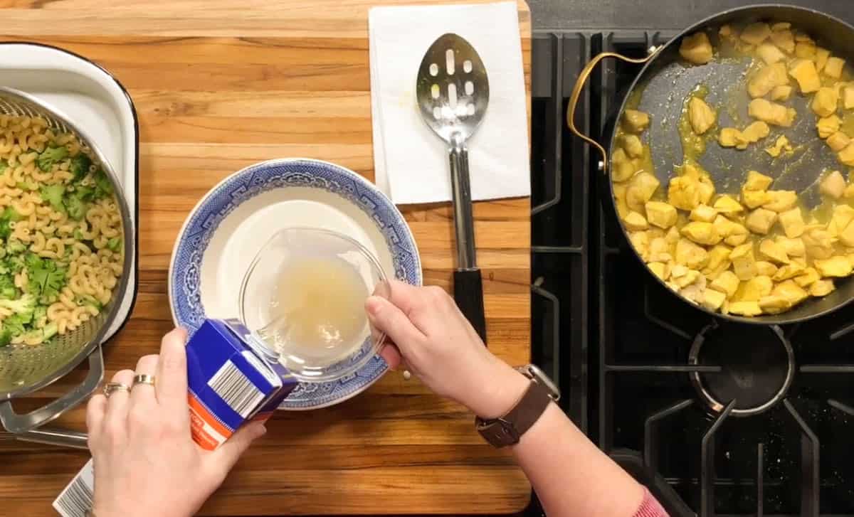 A person pours liquid from a measuring cup into a bowl of cream on a wooden counter. Nearby are a pan with cooked chicken pieces and a bowl of pasta with broccoli. A slotted spoon rests on a paper towel.