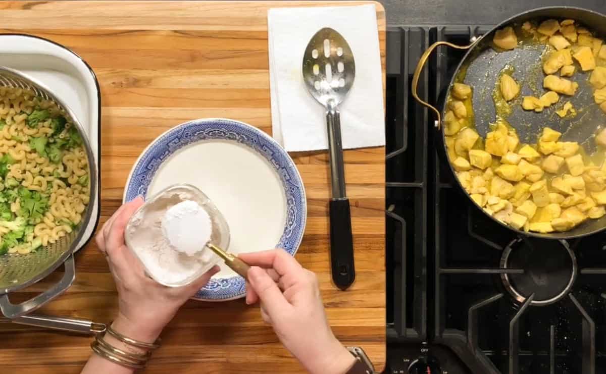 A person measures white powder from a jar over a bowl. Nearby are cooked pasta with greens, a slotted spoon on a napkin, and a skillet of yellow diced chicken on a stovetop.