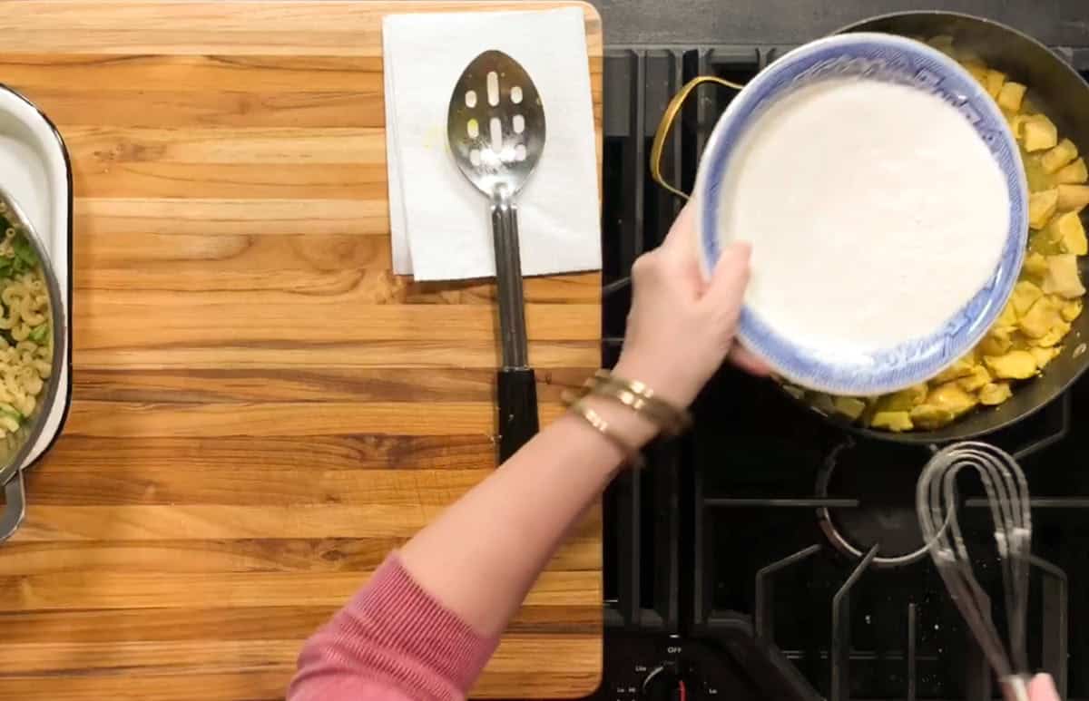 A person holds a bowl of white sauce over a pan with diced yellow ingredients on a stovetop. A whisk is visible in the pan, and a serving spoon rests on a napkin on a wooden countertop nearby.
