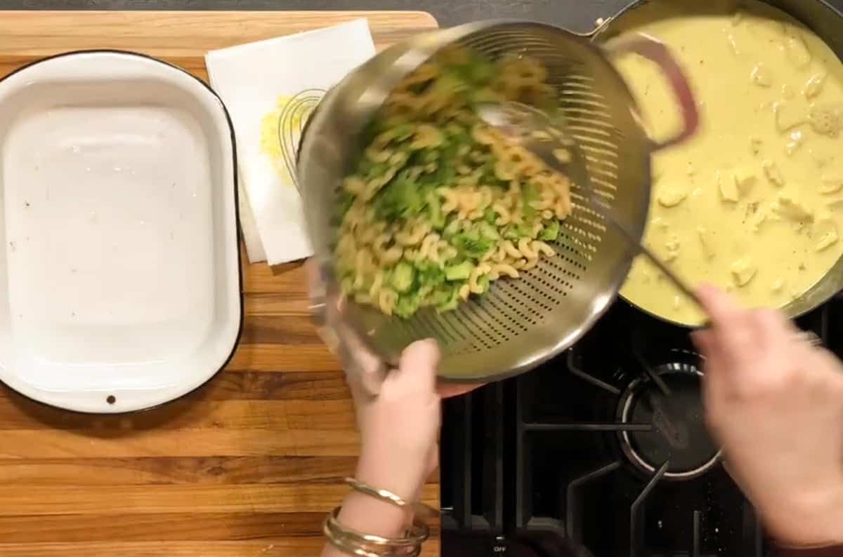 A person pours cooked pasta and green vegetables from a colander into a baking dish, with a pot of yellow sauce and cubed ingredients on the stove beside a wooden cutting board.