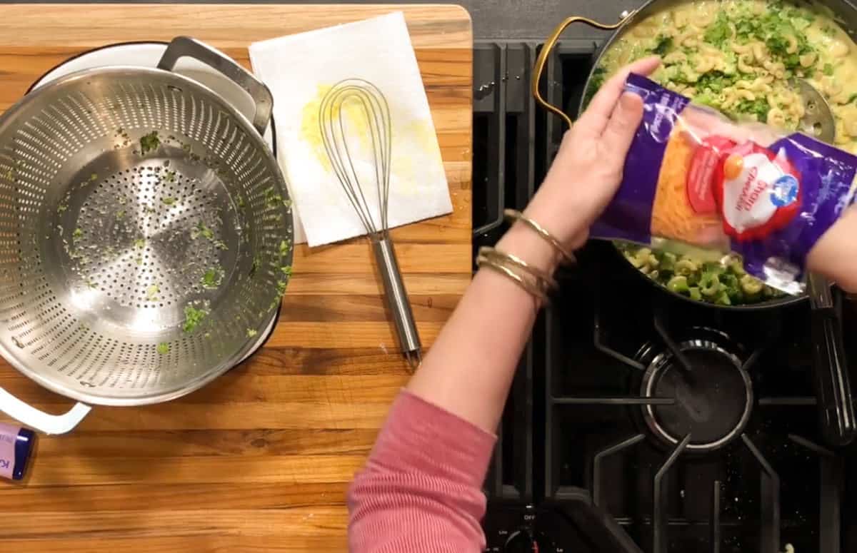 A person in a pink sweater adds shredded cheese from a bag into a pot of pasta and broccoli on a stove. A metal colander, whisk, and paper towel are on a wooden countertop nearby.