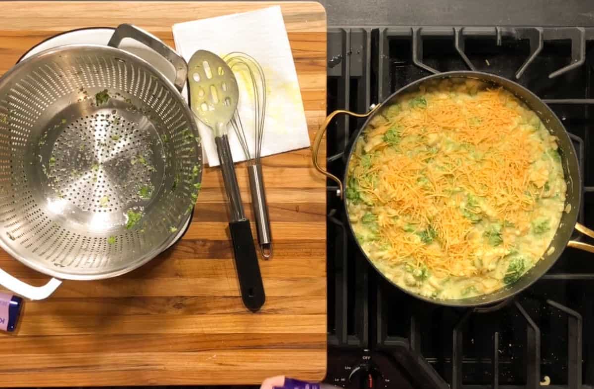 A metal colander with vegetable remnants sits on a wooden counter next to a slotted spoon, whisk, and paper towel. On the stove, a large pan contains a broccoli and cheese mixture.
