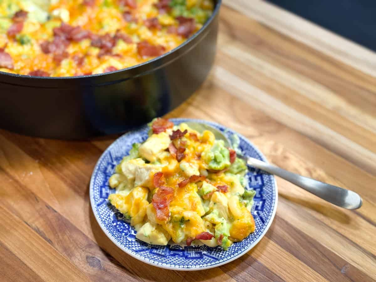 A blue patterned plate holds a serving of cheesy casserole with chunks of potato, broccoli, and bacon. A larger pot with more of the casserole is in the background, all on a wooden table with a spoon beside the plate.