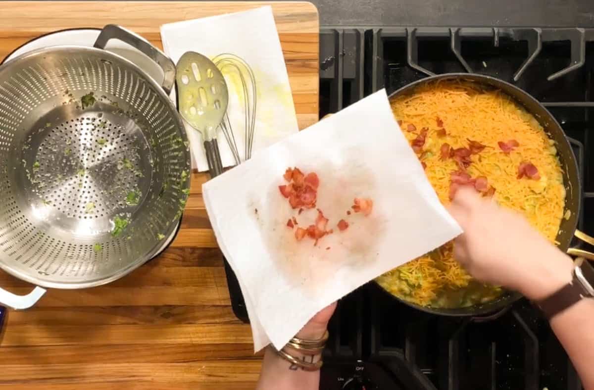 A person holds a paper towel with cooked bacon bits over a stove, preparing to add them to a pan of shredded cheese-topped food. A colander, utensils, and paper towels rest on a wooden counter nearby.