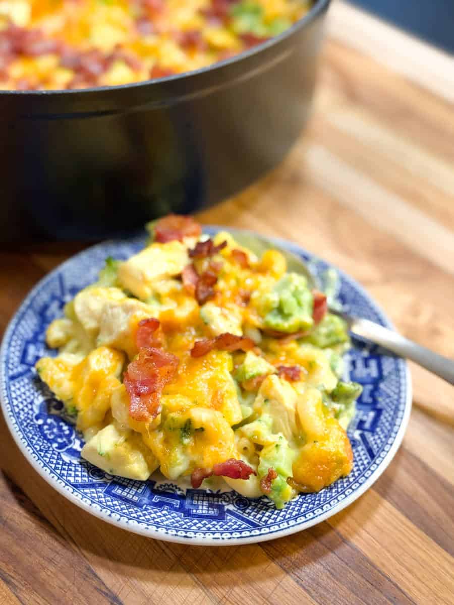 A blue and white plate holds a serving of cheesy casserole with visible broccoli, cauliflower, melted cheddar cheese, and bacon pieces. A fork rests beside the plate on a wooden surface, with a casserole dish in the background.