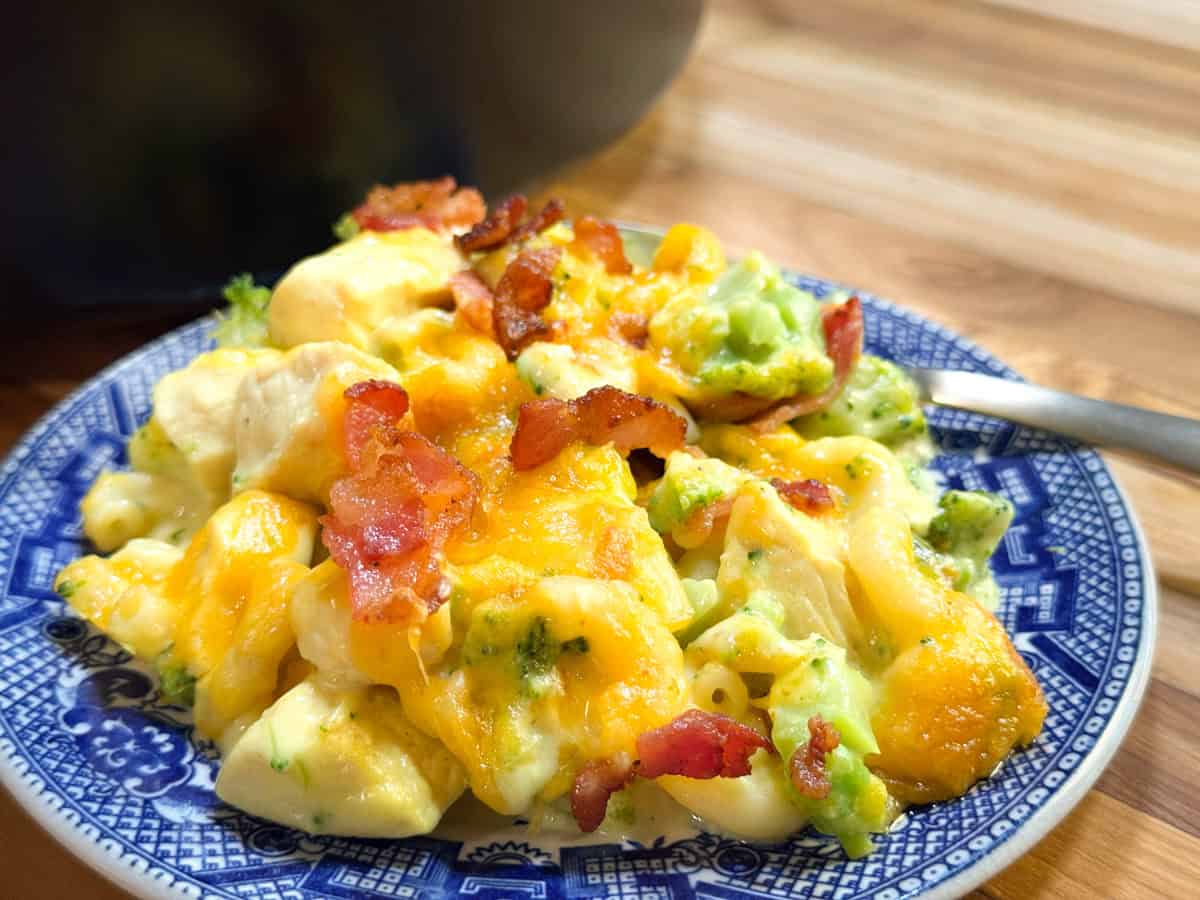 A blue and white plate holds a serving of macaroni and cheese with broccoli, topped with melted cheddar cheese and crispy bacon pieces, on a wooden table.