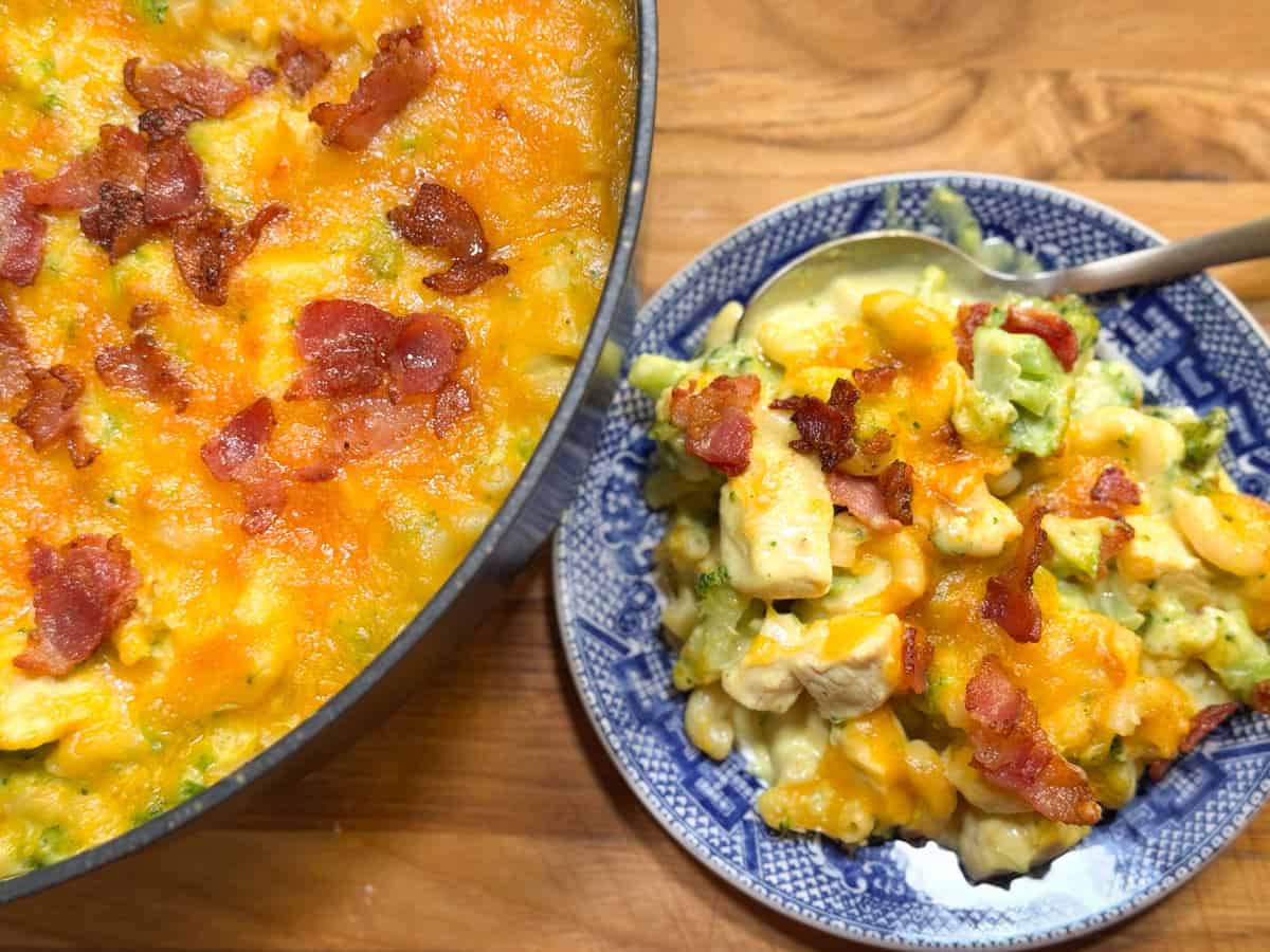 A casserole dish filled with cheesy baked pasta topped with pieces of bacon is next to a blue-patterned plate holding a serving of the same dish, which includes visible chunks of chicken and broccoli. A spoon is on the plate.