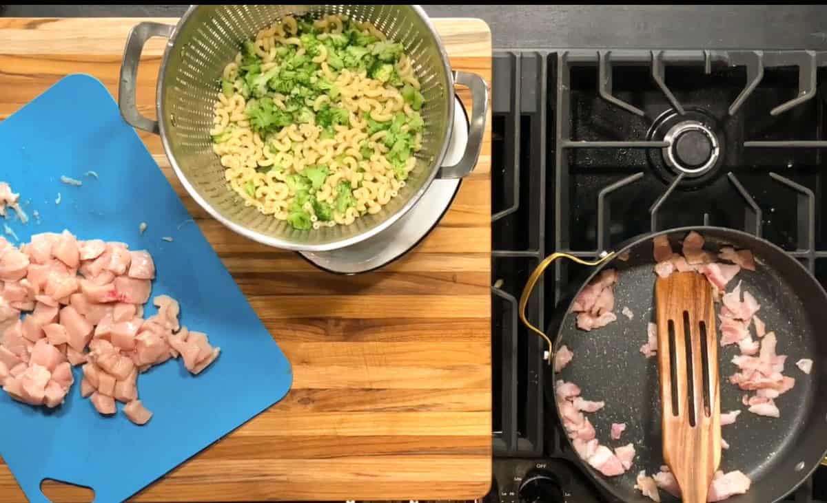 Chopped raw chicken on a blue cutting board, cooked elbow macaroni and broccoli in a colander over a pot, and diced bacon cooking in a skillet on a stove beside a wooden spoon.