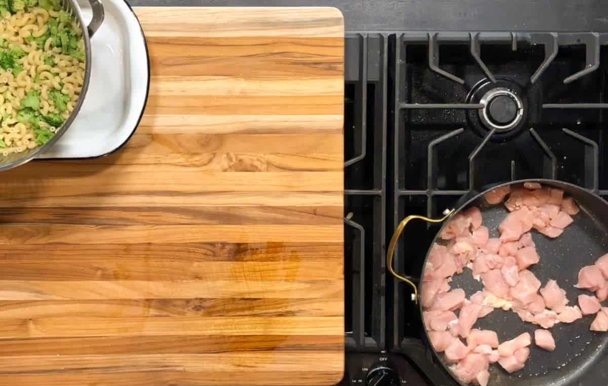 A pan with cubed raw chicken cooks on a stovetop next to a wooden cutting board. To the left, a pot contains cooked pasta and broccoli.