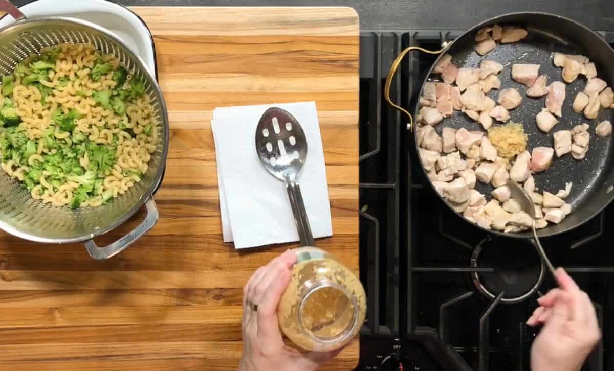 A person adds minced garlic to a frying pan with pieces of chicken on a stovetop. Next to the stove is a wooden cutting board holding a slotted spoon, napkin, and a colander of cooked pasta with broccoli.