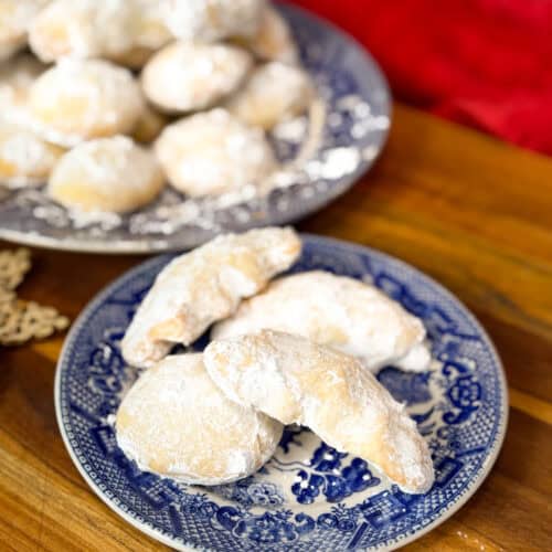A blue and white patterned plate holds three powdered sugar-covered cookies, with more cookies on a similar plate in the background. The plates are on a wooden surface.