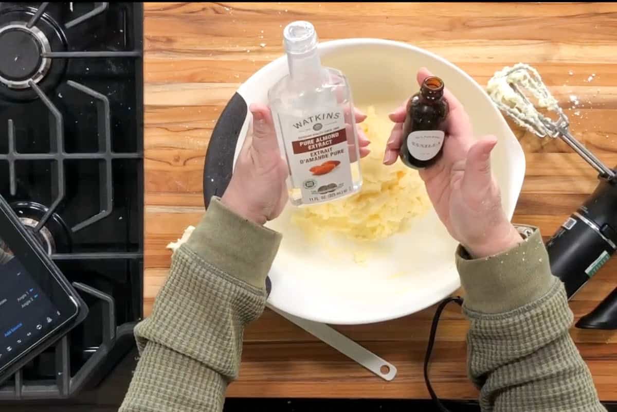 A person holds bottles of almond and vanilla extracts above a white mixing bowl with creamed ingredients on a wooden countertop, next to a hand mixer and a stovetop.