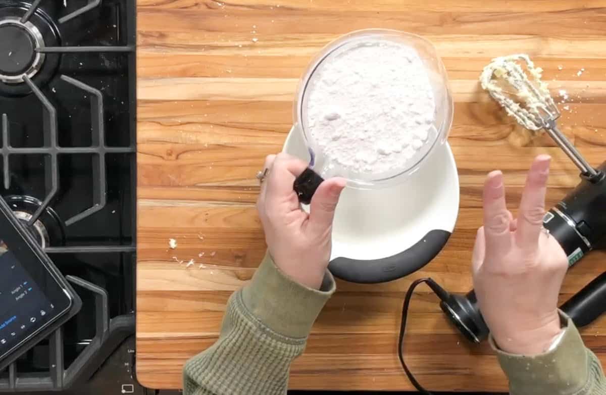 A person holds a measuring cup filled with flour over a kitchen scale on a wooden countertop. A hand mixer with batter on the beaters and a digital tablet are also visible nearby.