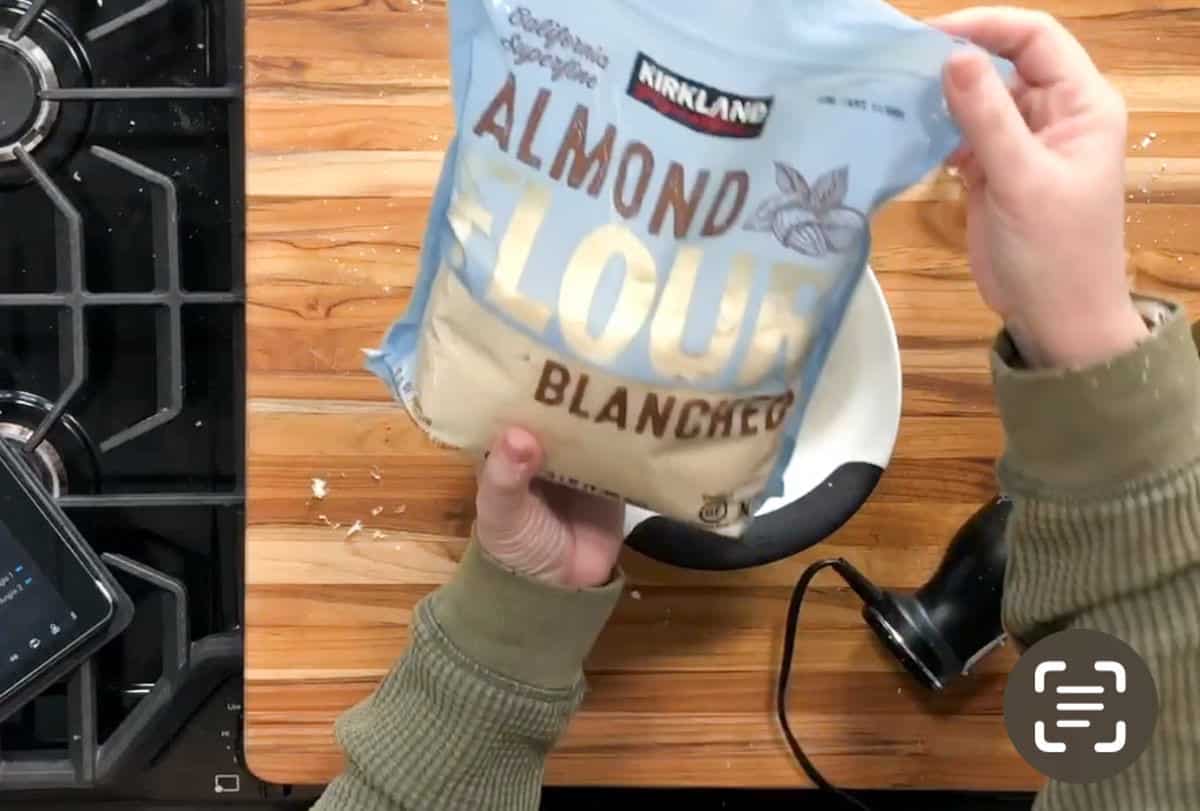 A person holds a bag of Kirkland brand blanched almond flour over a mixing bowl on a wooden countertop. A mixer, a stovetop, and a digital device are also visible nearby.