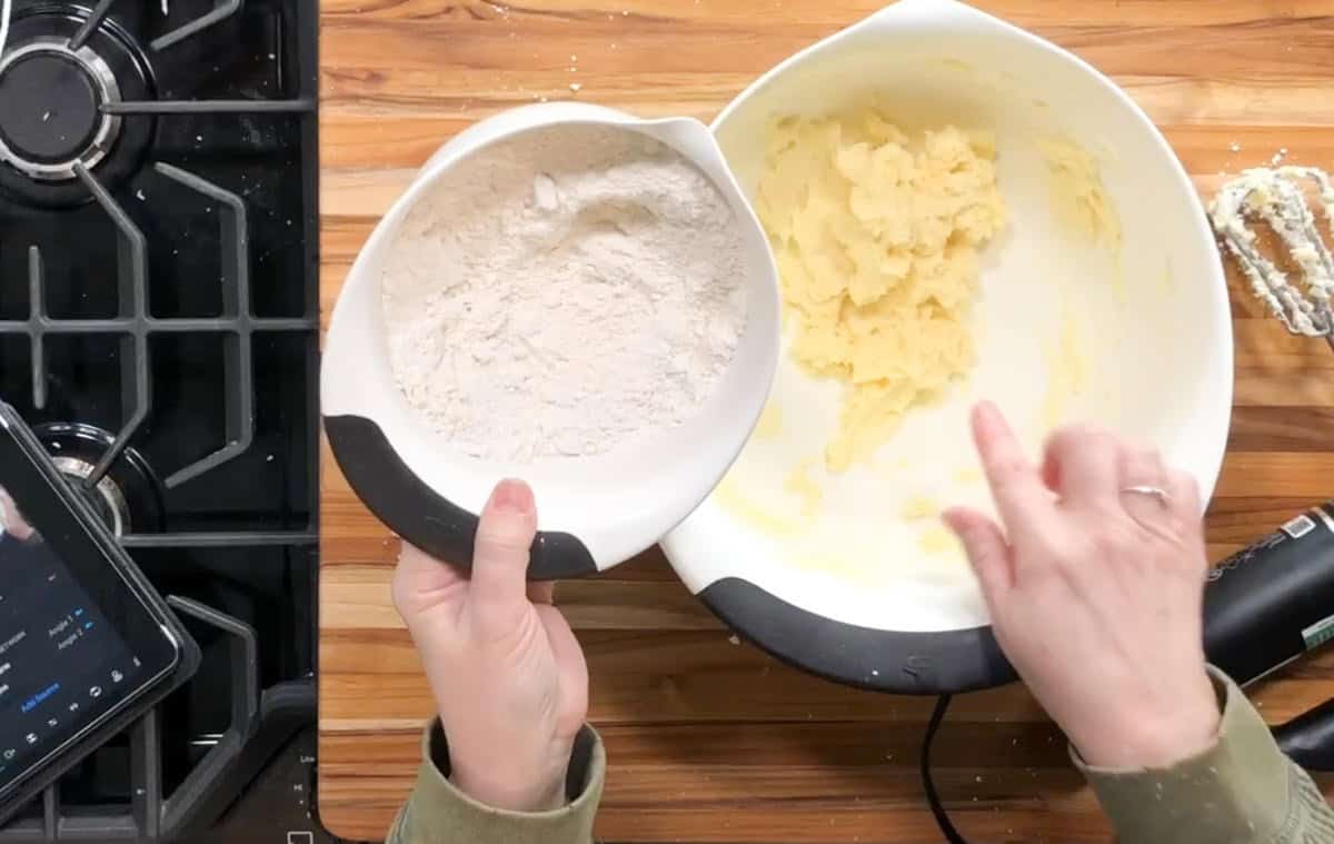 A person holds a bowl of flour over a larger bowl with creamed butter and sugar, preparing to mix ingredients on a wooden countertop next to a gas stove, a tablet, and a hand mixer.