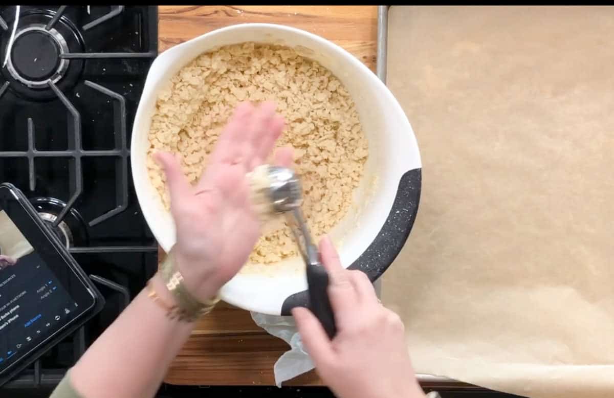 A person scoops crumbly dough from a white bowl onto their hand using a metal scoop, next to a parchment-lined baking sheet on a wooden counter near a stove.