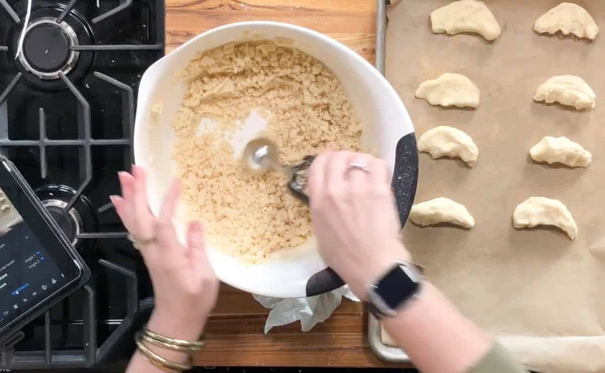 A person mixes dough in a white bowl next to a baking sheet with unbaked pastries shaped like half-moons; a stove and tablet are visible on the left side of the image.