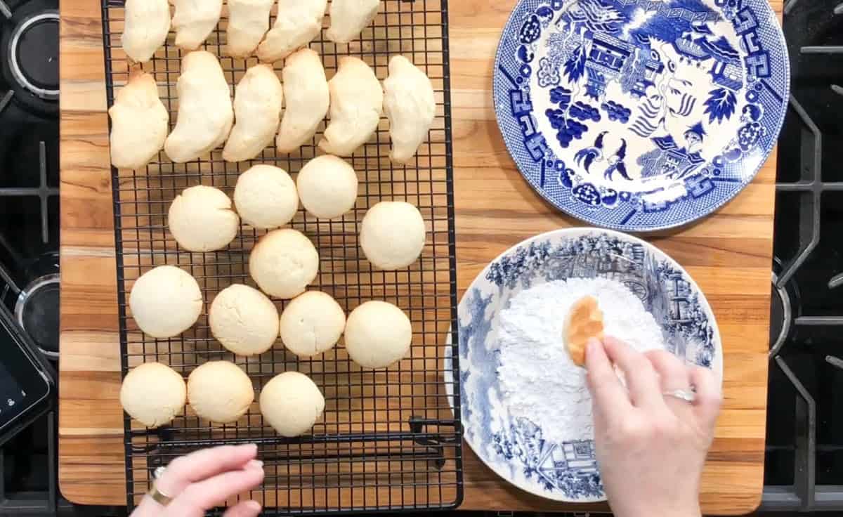 A hand dips a round cookie into a bowl of powdered sugar. Several cookies cool on a wire rack beside an empty blue-and-white patterned plate. The setup is on a wooden surface next to a stovetop.