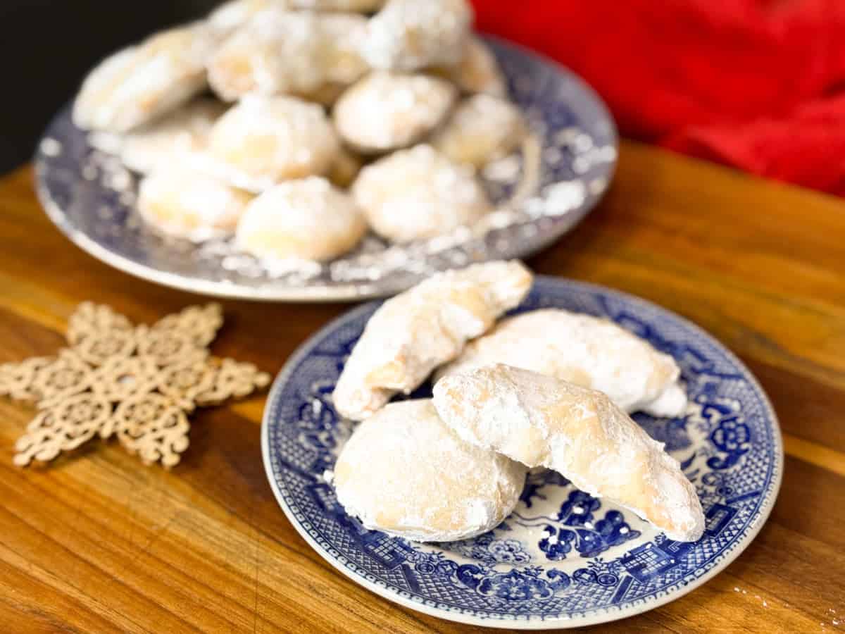 Two blue and white plates hold crescent-shaped and round cookies dusted with powdered sugar on a wooden surface. A wooden snowflake ornament and a red cloth are nearby.