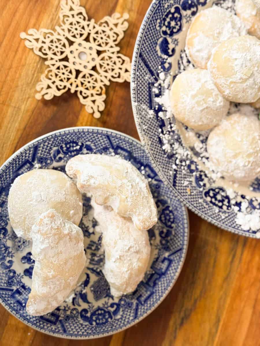 Two blue and white patterned plates on a wooden surface hold crescent-shaped and round cookies dusted with powdered sugar. A wooden snowflake ornament is placed next to the plates.