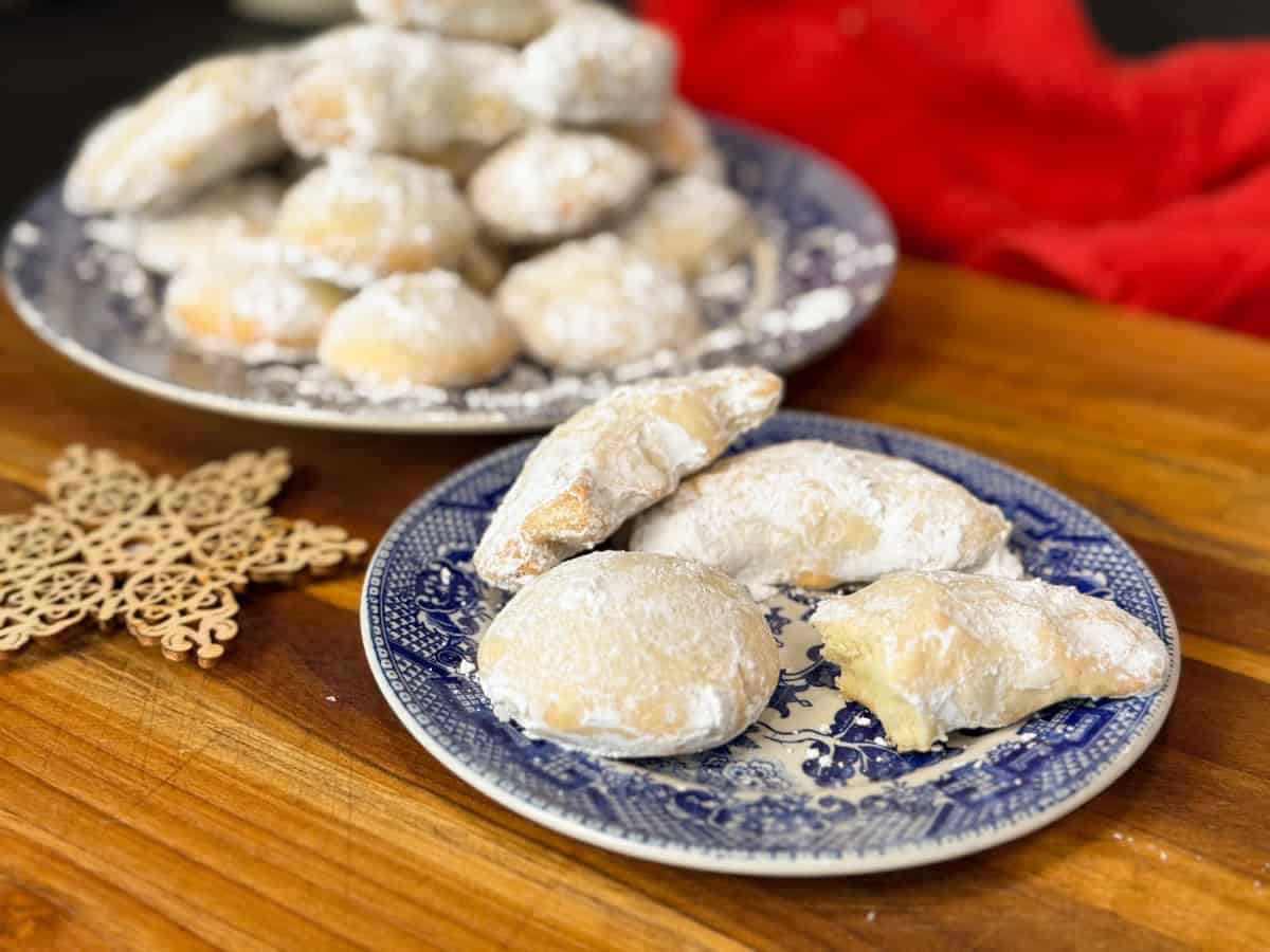Two blue and white patterned plates hold crescent-shaped and round cookies dusted with powdered sugar, arranged on a wooden surface with a decorative snowflake and a red cloth in the background.