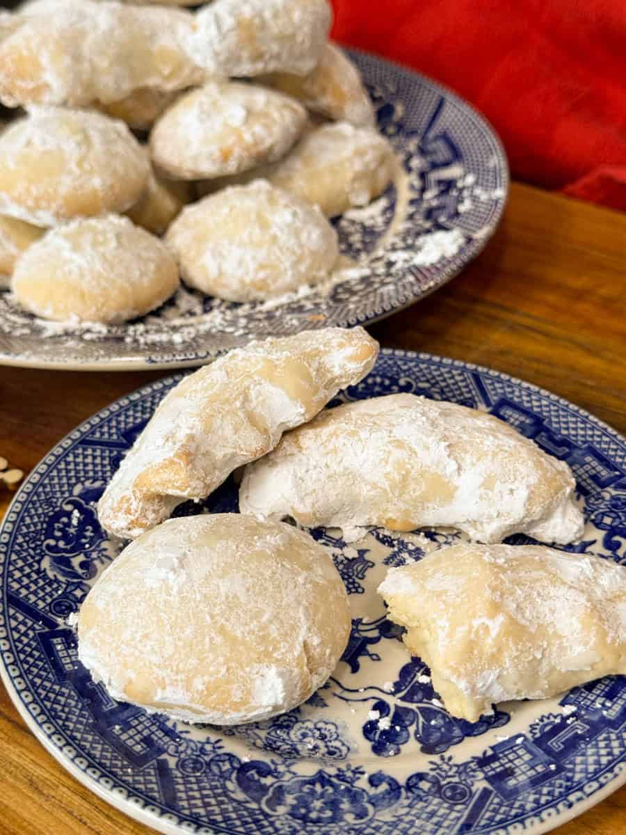 Powdered sugar-coated cookies are arranged on blue and white patterned plates. Some cookies are whole, while others are partially broken, showing a soft, light interior. A red cloth is visible in the background.